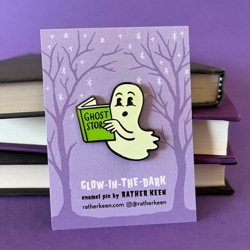 Enamel pin in the shape of a cartoon ghost reading a book of spooky stories