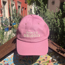 Vamos a la Biblioteca hat in dusty-rose pink and off-white embroidery on the front, posed outdoors with library in the background