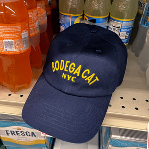 Navy blue dad hat with Bodega Cat NYC embroidered in yellow thread sitting on a bodega shelf