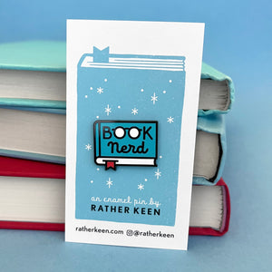 enamel pin shaped like a book with the words Book Nerd on the cover.