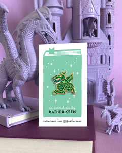 The green Book Wyrm dragon pin on its display card held by a purple toy dragon