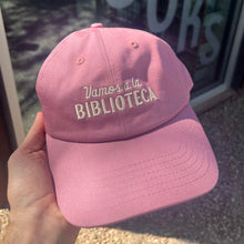 A dusty-rose pink "dad hat" held in hand, embroidered with "Vamos a la Biblioteca"